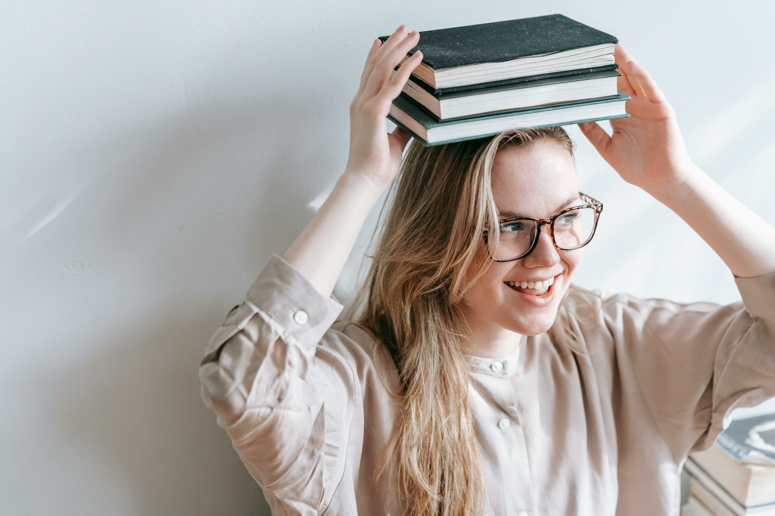 Une jeune femme souriante, portant des lunettes, tenant une pile de trois livres sur sa tête avec les mains.