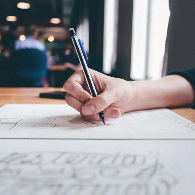 Une main tenant un crayon, écrivant ou dessinant sur une feuille de papier posée sur une table en bois.