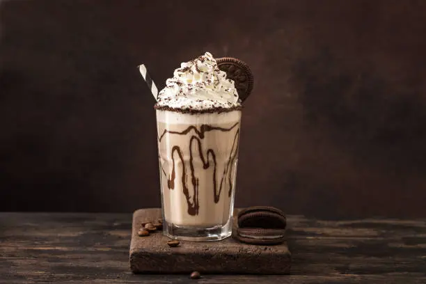 Un grand verre de milkshake chocolat-café garni de crème fouettée, de sauce chocolat, de brisures et d'un biscuit Oreo, posé sur une planche en bois.