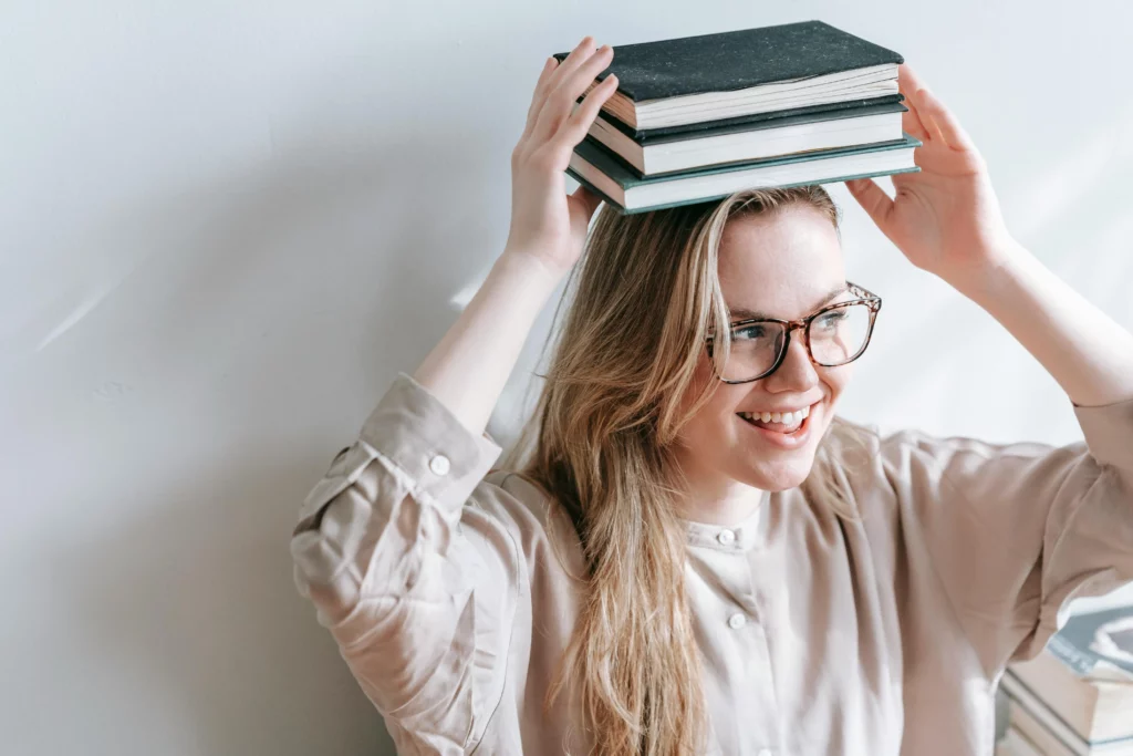 Une jeune femme souriante, portant des lunettes, tenant une pile de trois livres sur sa tête avec les mains.